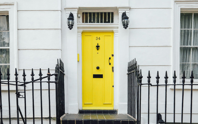 A clean white wall, with a black metal fence, in the centre sits a bright contrasting yellow front door.