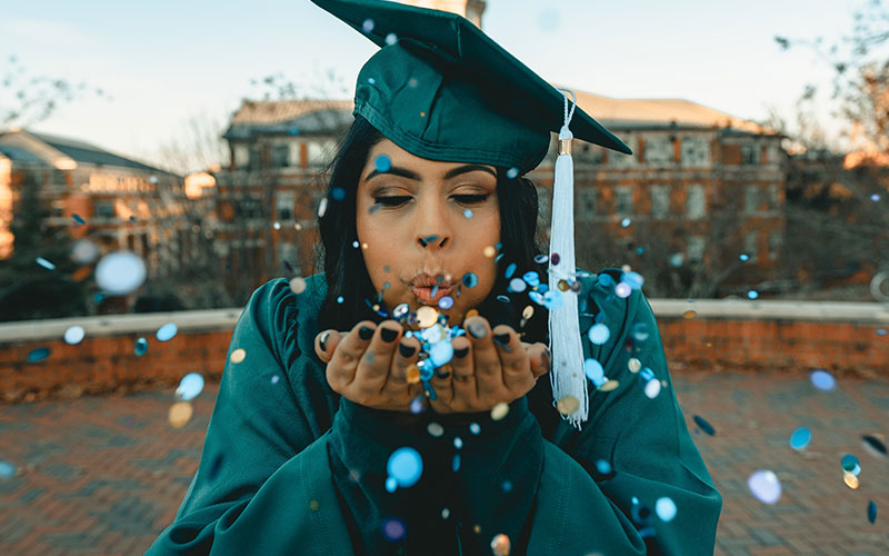 A woman wearing her graduation gown and cap, blowing glitter towards the camera.
