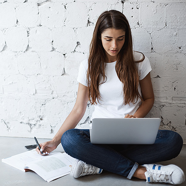 Indoor portrait of smart busy woman, making notes while leaning on wall and sitting on floor with laptop on crossed feet. Girl has habit to work or study everywhere but not at table. Study and work
