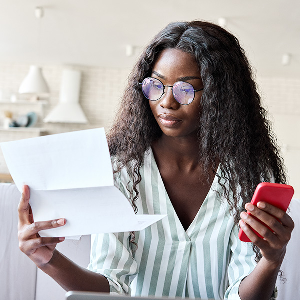 Young intelligent serious African black businesswoman in glasses sits on sofa reading official legal paper letter using mobile banking application on cell mobile smartphone at modern home office.