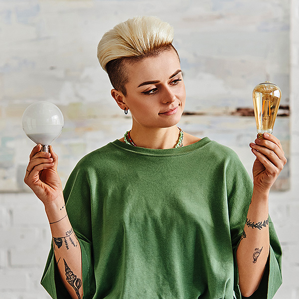 stylish tattooed woman in casual clothes, with trendy hairstyle comparing different energy saving light bulbs in modern living room, sustainable lifestyle and environmentally conscious concept