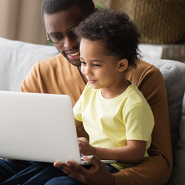 African father holds little son on lap family sitting on sofa at home with notebook, looking at screen watching cartoon educational videos, make video call, having fun enjoy free time together concept