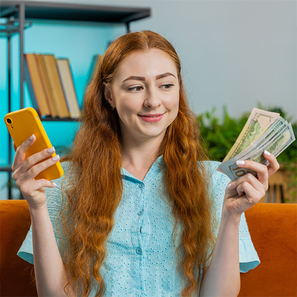 Planning family budget. Smiling young redhead woman counting money cash, use smartphone calculate domestic bills at home room. Girl satisfied of income and saves money for planned vacation, gifts