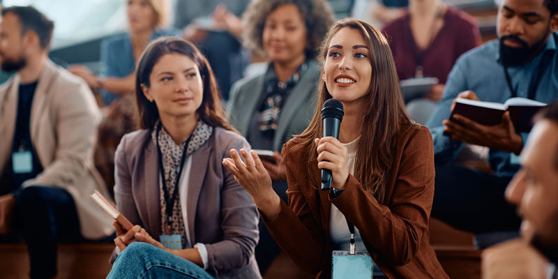 A happy young lady talking into a microphone at a large meeting in a conference centre.